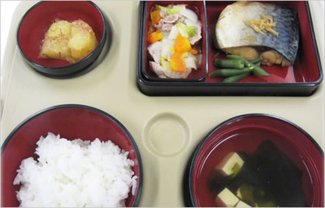 介護施設の食事提供。赤い食器に盛られた白米、汁物、弁当箱の各種料理